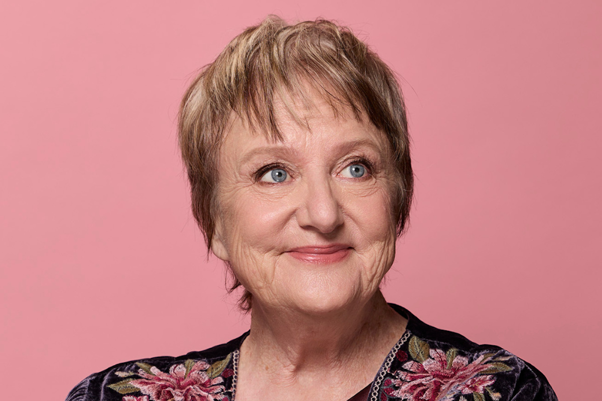Studio portrait of Australian comedian and actress Denise Scott smiling against a solid pink background, wearing an embroidered floral dark top.